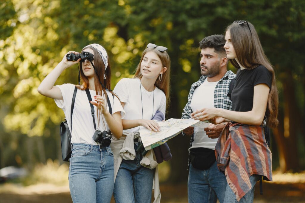 A diverse group of friends use a map and binoculars in a park, enjoying a sunny day outdoors.