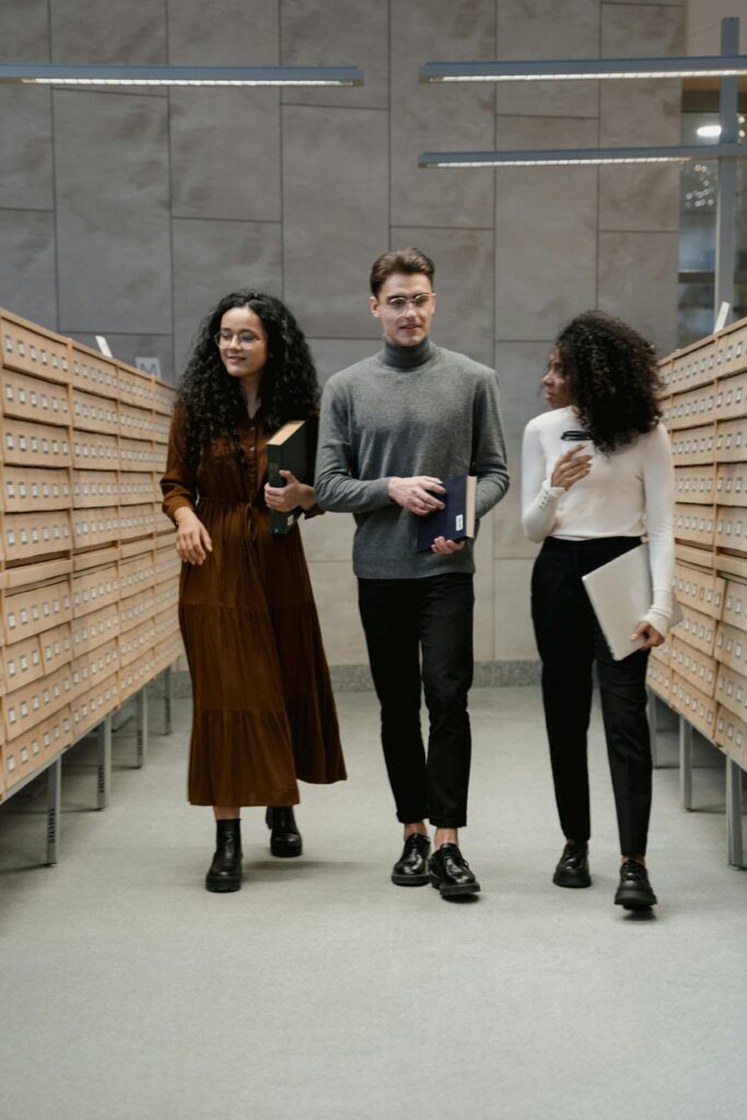 pexels-photo-6549583-6549583 Three diverse students walking in a library, holding books, discussing academic topics.